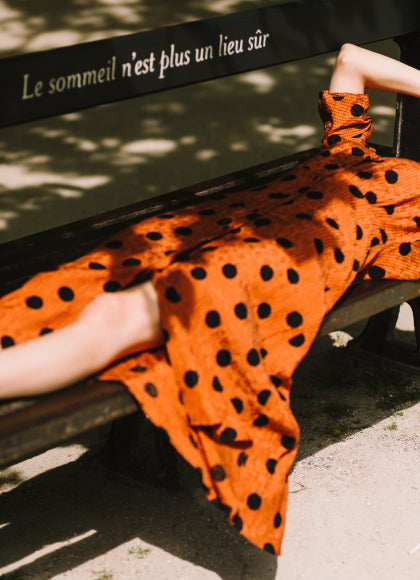 Person wearing an orange dress with black polka dots sitting on a bench with text in the background.